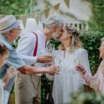 Mature bride and groom kissing at wedding reception with their family, outside in the backyard.